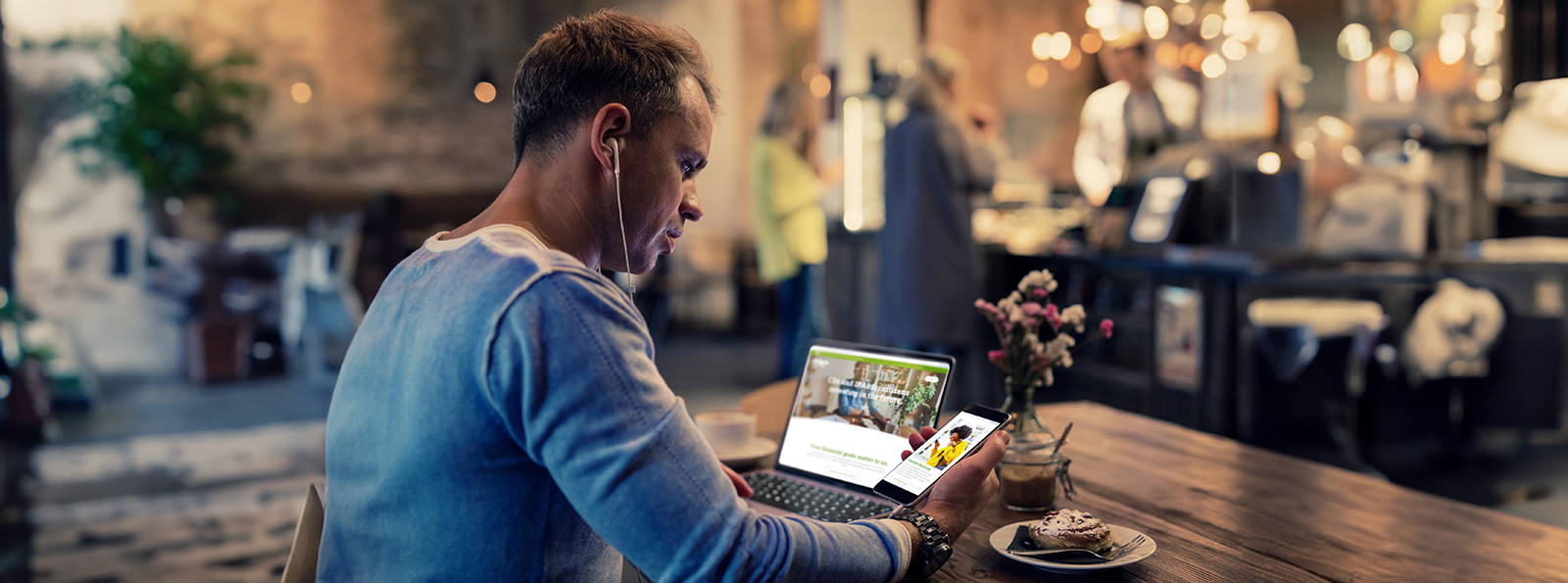 A man wearing earbuds is seated in a cafe  looking at his smartphone and his laptop