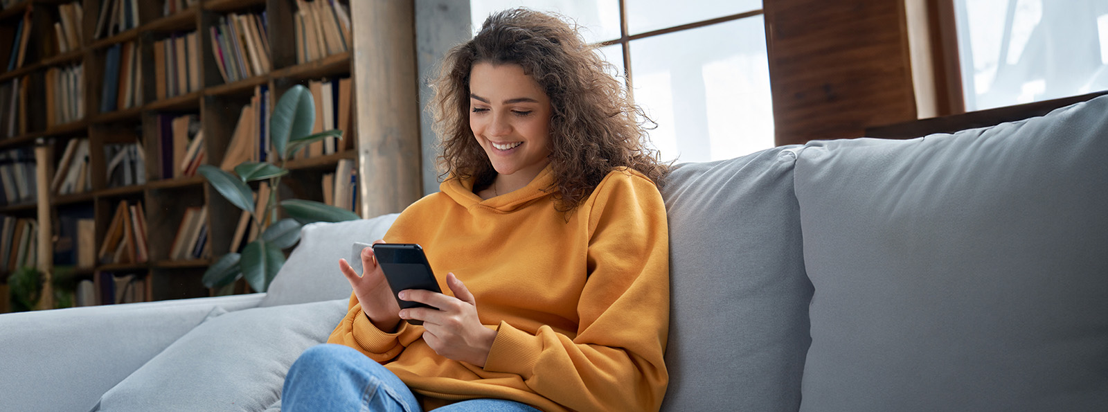 Young woman is sitting on a couch holding her smart phone and scrolling