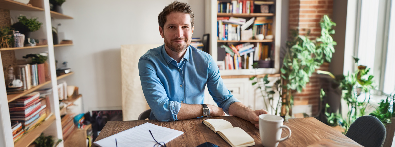 A man sits at his home office with a note pad and coffee on his desk as he smiles at the camera