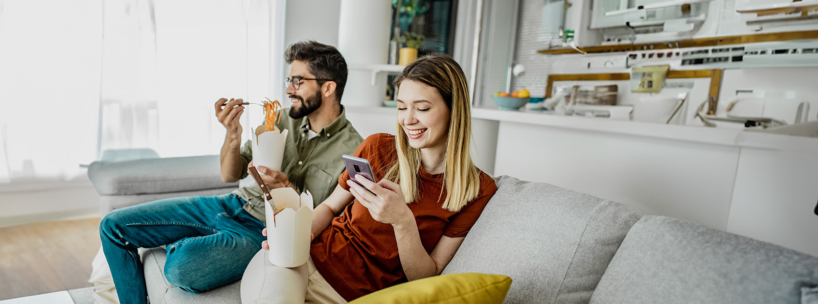 A guy and girl are sitting on the couch together eating takeout food