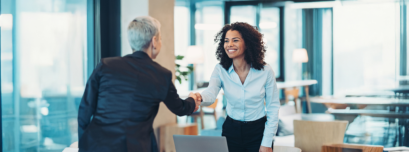 two professionally-dressed women shake hands in a modern corporate setting