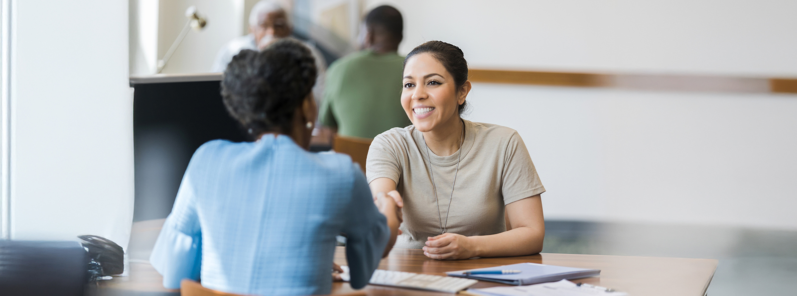 to professionally dressed women seated across the table from one another shaking hands and smiling