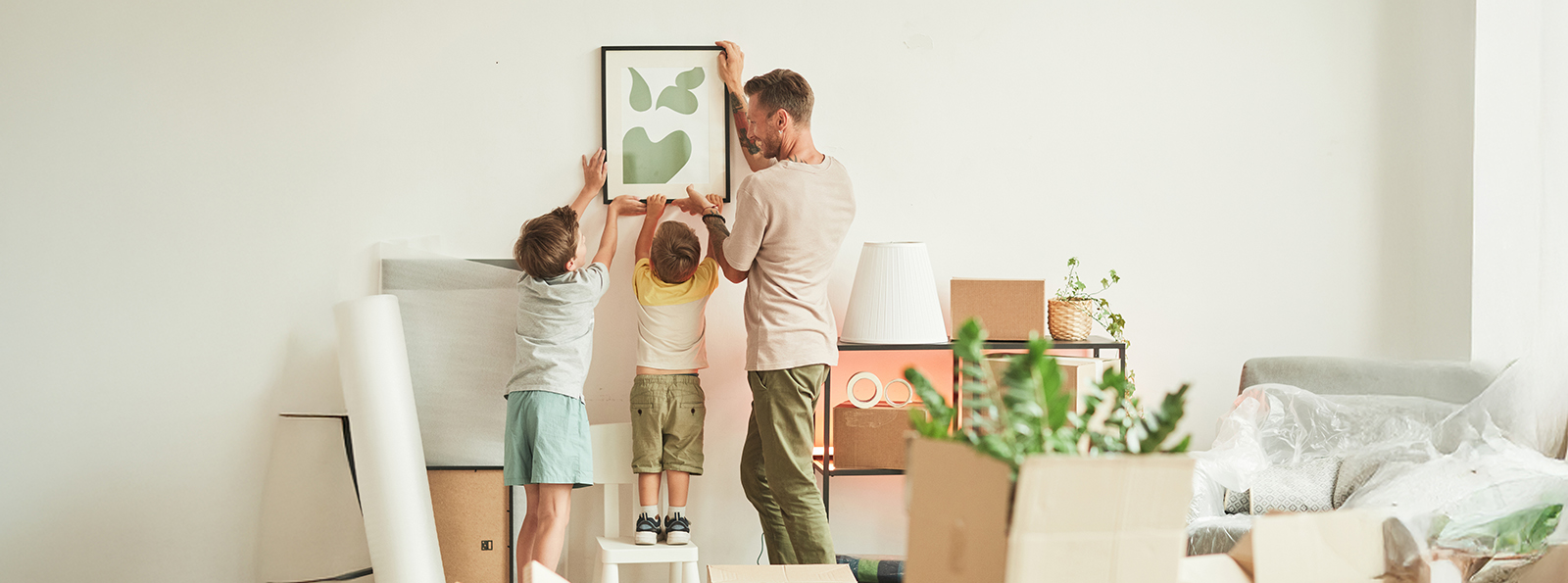 a father and two of his children hanging a framed painting on an interior wall of their house