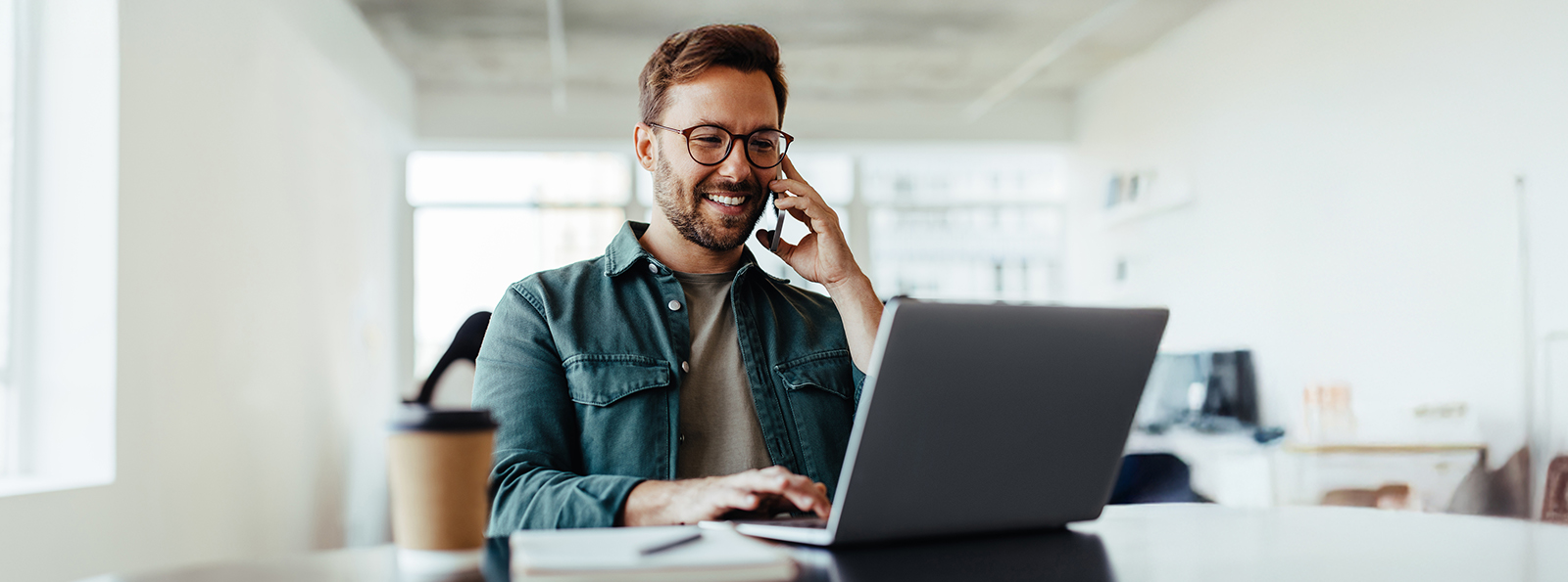 A man in his late 30's wearing glasses uses a telephone while reviewing information on his laptop